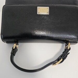 K&ouml;nnte beinhalten: Eine schwarze, gl&auml;nzende Handtasche mit rechteckiger Form und einem oberen Griff. Die Tasche hat goldfarbene Hardware und ein kleines, rechteckiges Metallschild auf der Vorderseite.