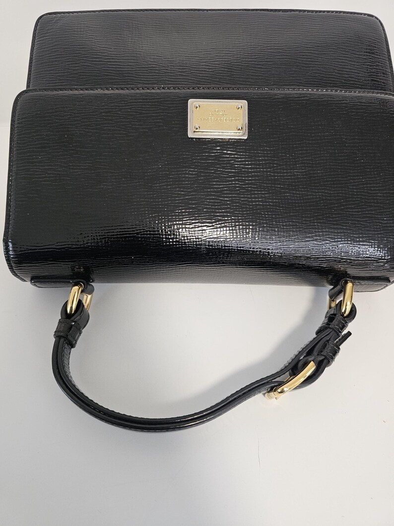 K&ouml;nnte beinhalten: Eine schwarze, gl&auml;nzende Handtasche mit rechteckiger Form und einem oberen Griff. Die Tasche hat goldfarbene Hardware und ein kleines, rechteckiges Metallschild auf der Vorderseite.