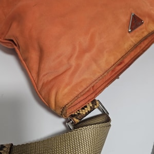 Peut inclure: Sac &agrave; bandouli&egrave;re en nylon orange avec un d&eacute;tail en peau de serpent et une boucle argent&eacute;e. Le sac pr&eacute;sente un logo triangulaire en m&eacute;tal. La bandouli&egrave;re est de couleur beige.