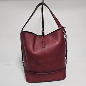 以下が含まれることがあります： シルバーの鍵とチャームが付いた、ワインレッドの革製のバケットバッグ。バッグには、トップハンドルが1つと取り外し可能なショルダーストラップが付いています。