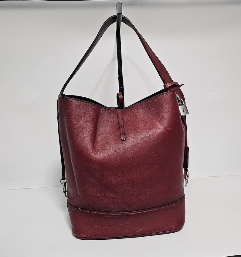 以下が含まれることがあります： シルバーの鍵とチャームが付いた、ワインレッドの革製のバケットバッグ。バッグには、トップハンドルが1つと取り外し可能なショルダーストラップが付いています。