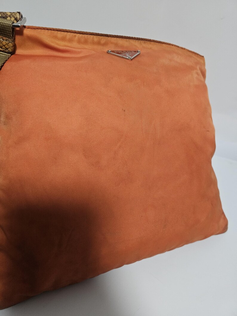 Peut inclure: Un sac &agrave; main orange avec un logo triangulaire argent&eacute;. Le sac a une bandouli&egrave;re en tissu avec un motif peau de serpent. Le sac est fait d'une mati&egrave;re douce, possiblement en nylon.