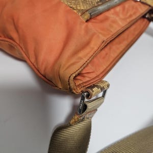 Peut inclure: Sac &agrave; bandouli&egrave;re orange et motif peau de serpent, avec une fermeture &eacute;clair argent&eacute;e. Il poss&egrave;de une bandouli&egrave;re tiss&eacute;e et une boucle m&eacute;tallique avec la marque "PRADA". Le sac est fait de plusieurs mat&eacute;riaux.