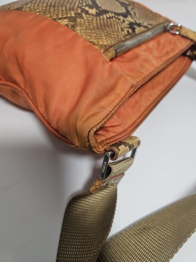Peut inclure: Sac &agrave; bandouli&egrave;re orange et motif peau de serpent, avec une fermeture &eacute;clair argent&eacute;e. Il poss&egrave;de une bandouli&egrave;re tiss&eacute;e et une boucle m&eacute;tallique avec la marque "PRADA". Le sac est fait de plusieurs mat&eacute;riaux.