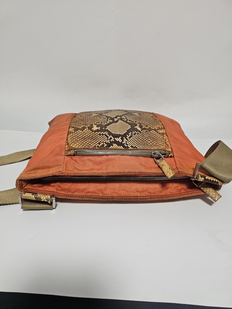 Peut inclure: Un sac bandouli&egrave;re orange avec un panneau en peau de serpent et une fermeture &eacute;clair. Le sac a une sangle beige et des accents en peau de serpent sur les attaches de la sangle. Le sac est en mati&egrave;re de type nylon.