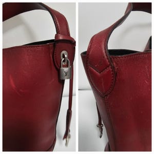 以下が含まれることがあります： 銀色の鍵と錠前が付いた赤い革のバケットバッグ。バッグはトップハンドルとショルダーストラップが付いています。