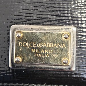 K&ouml;nnte beinhalten: Nahaufnahme eines schwarzen Lederaccessoires mit strukturierter Oberfl&auml;che. Eine rechteckige Metallplatte mit den Worten "DOLCE & GABBANA MILANO ITALIA" ist angebracht. Die Platte hat eine goldfarbene Mitte und silberfarbene R&auml;nder.