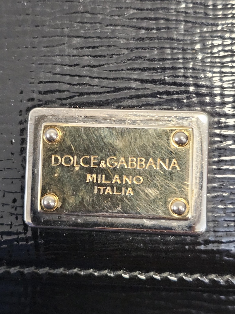 K&ouml;nnte beinhalten: Nahaufnahme eines schwarzen Lederaccessoires mit strukturierter Oberfl&auml;che. Eine rechteckige Metallplatte mit den Worten "DOLCE & GABBANA MILANO ITALIA" ist angebracht. Die Platte hat eine goldfarbene Mitte und silberfarbene R&auml;nder.