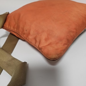 Peut inclure: Un sac carr&eacute; orange avec une sangle beige. Le sac pr&eacute;sente des signes d'usure, avec une d&eacute;coloration sur le tissu. La sangle est en mat&eacute;riau tiss&eacute;. Le sac semble &ecirc;tre fait d'un tissu durable, peut-&ecirc;tre du nylon. Id&eacute;al pour transporter des objets.
