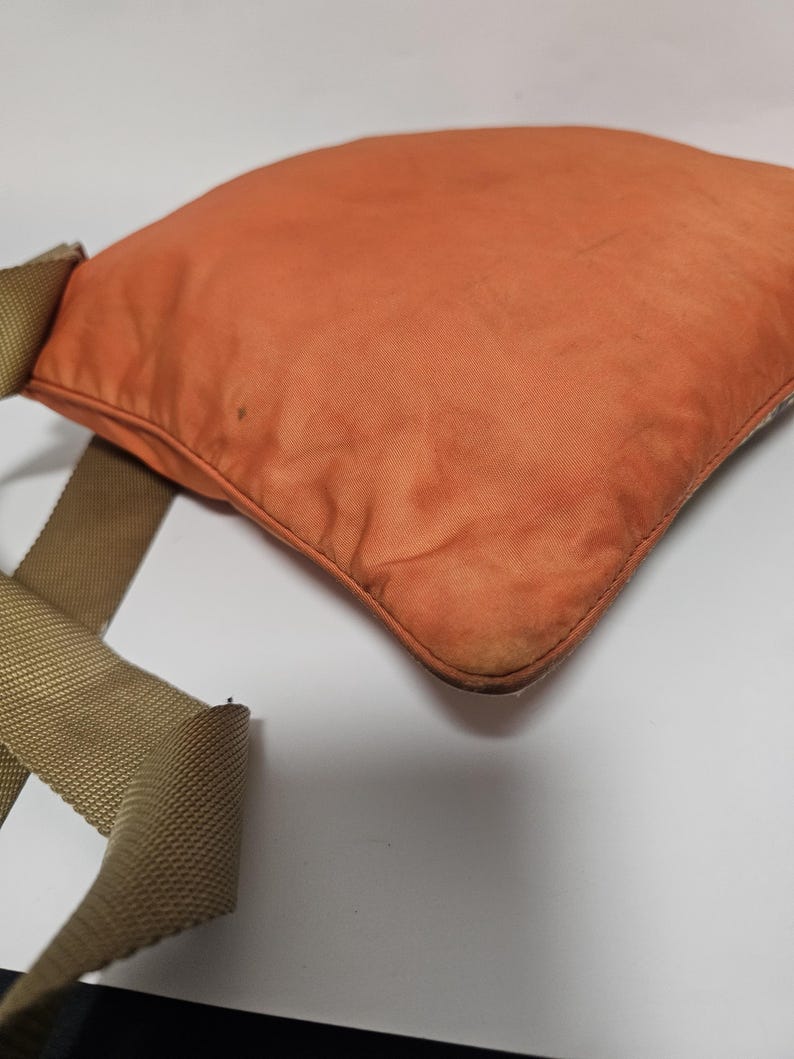 Peut inclure: Un sac carr&eacute; orange avec une sangle beige. Le sac pr&eacute;sente des signes d'usure, avec une d&eacute;coloration sur le tissu. La sangle est en mat&eacute;riau tiss&eacute;. Le sac semble &ecirc;tre fait d'un tissu durable, peut-&ecirc;tre du nylon. Id&eacute;al pour transporter des objets.
