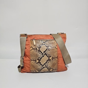 以下が含まれることがあります： 前面にヘビ革のパネルが付いたオレンジ色のクロスボディバッグ。バッグには、長さ調節可能なショルダーストラップとジッパー付きポケットが付いています。バッグは耐久性のある素材で作られています。