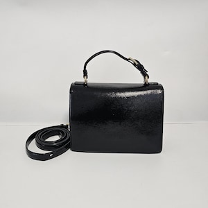 K&ouml;nnte beinhalten: Eine schwarze, rechteckige Handtasche mit einem oberen Griff und einem abnehmbaren Schulterriemen. Die Tasche hat eine strukturierte Oberfl&auml;che und goldfarbene Hardware. Der Riemen ist auf der linken Seite der Tasche aufgerollt.