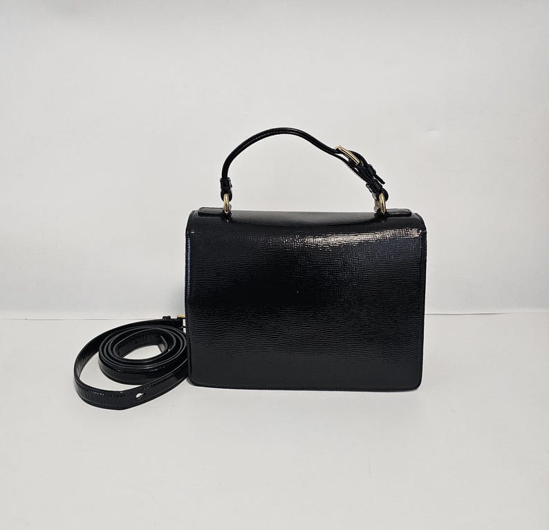 K&ouml;nnte beinhalten: Eine schwarze, rechteckige Handtasche mit einem oberen Griff und einem abnehmbaren Schulterriemen. Die Tasche hat eine strukturierte Oberfl&auml;che und goldfarbene Hardware. Der Riemen ist auf der linken Seite der Tasche aufgerollt.