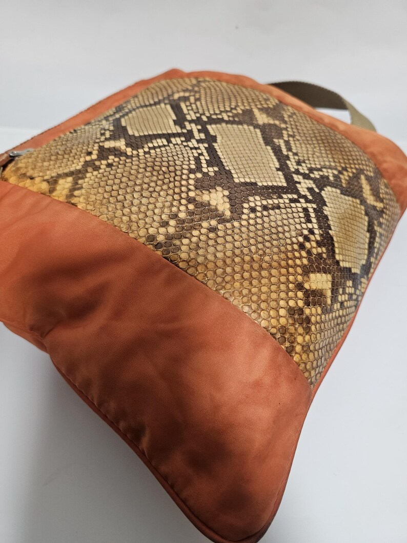 Peut inclure: Coussin carr&eacute; avec un panneau central en peau de serpent et une bordure en tissu couleur rouille. Le motif en peau de serpent pr&eacute;sente des nuances de marron, de beige et de cr&egrave;me. Une sangle verte est attach&eacute;e.