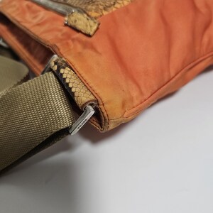 Peut inclure: Gros plan sur un sac bandouli&egrave;re Prada orange avec un rabat et des d&eacute;tails en peau de serpent. Le sac a une bandouli&egrave;re en toile beige et une fermeture &eacute;clair argent&eacute;e. Le logo Prada est visible sur le c&ocirc;t&eacute;.