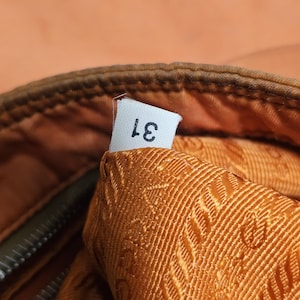 Peut inclure: Gros plan sur une section d'un sac, avec une &eacute;tiquette blanche portant le num&eacute;ro "31" imprim&eacute; en noir. L'int&eacute;rieur du sac est en tissu textur&eacute; orange dor&eacute;. L'ext&eacute;rieur est de couleur marron fonc&eacute;.