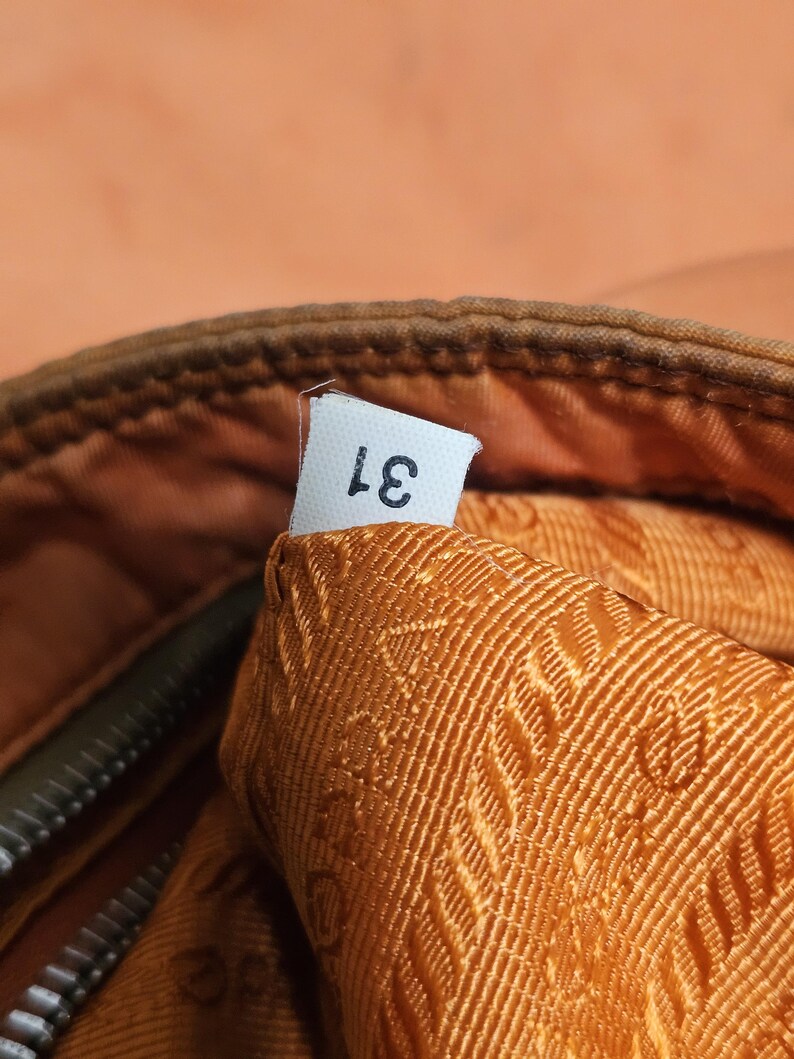 Peut inclure: Gros plan sur une section d'un sac, avec une &eacute;tiquette blanche portant le num&eacute;ro "31" imprim&eacute; en noir. L'int&eacute;rieur du sac est en tissu textur&eacute; orange dor&eacute;. L'ext&eacute;rieur est de couleur marron fonc&eacute;.