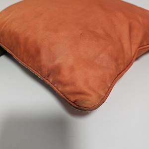 Peut inclure: Un coussin carr&eacute; orange avec un tissu textur&eacute;. Le coussin a une couture visible sur les bords et pr&eacute;sente des signes d'usure. La couleur est une teinte chaude et terreuse.