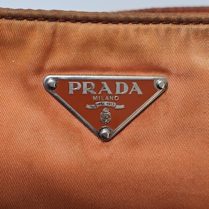 Peut inclure: Gros plan sur un sac &agrave; main Prada orange br&ucirc;l&eacute;. Le sac pr&eacute;sente un logo triangulaire en m&eacute;tal avec le nom de la marque et "MILANO DAL 1913" en lettres argent&eacute;es. Le mat&eacute;riau du sac a un l&eacute;ger &eacute;clat et les bords sont brun fonc&eacute;.