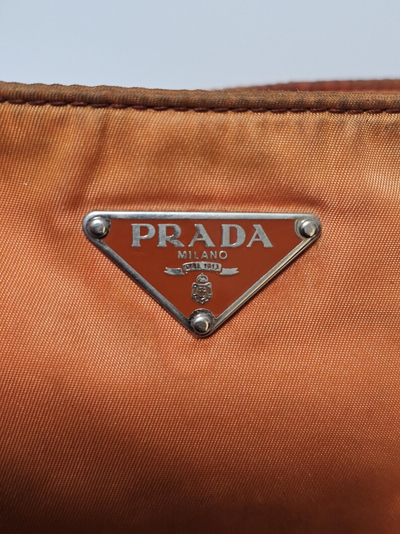 Peut inclure: Gros plan sur un sac &agrave; main Prada orange br&ucirc;l&eacute;. Le sac pr&eacute;sente un logo triangulaire en m&eacute;tal avec le nom de la marque et "MILANO DAL 1913" en lettres argent&eacute;es. Le mat&eacute;riau du sac a un l&eacute;ger &eacute;clat et les bords sont brun fonc&eacute;.