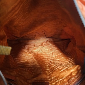 Peut inclure: Vue int&eacute;rieure d'un sac &agrave; main orange. La doublure est d'une nuance plus claire d'orange avec un motif r&eacute;p&eacute;titif. Une petite &eacute;tiquette m&eacute;tallique rectangulaire avec le nom de la marque est visible en bas. L'ext&eacute;rieur du sac est orange fonc&eacute;.