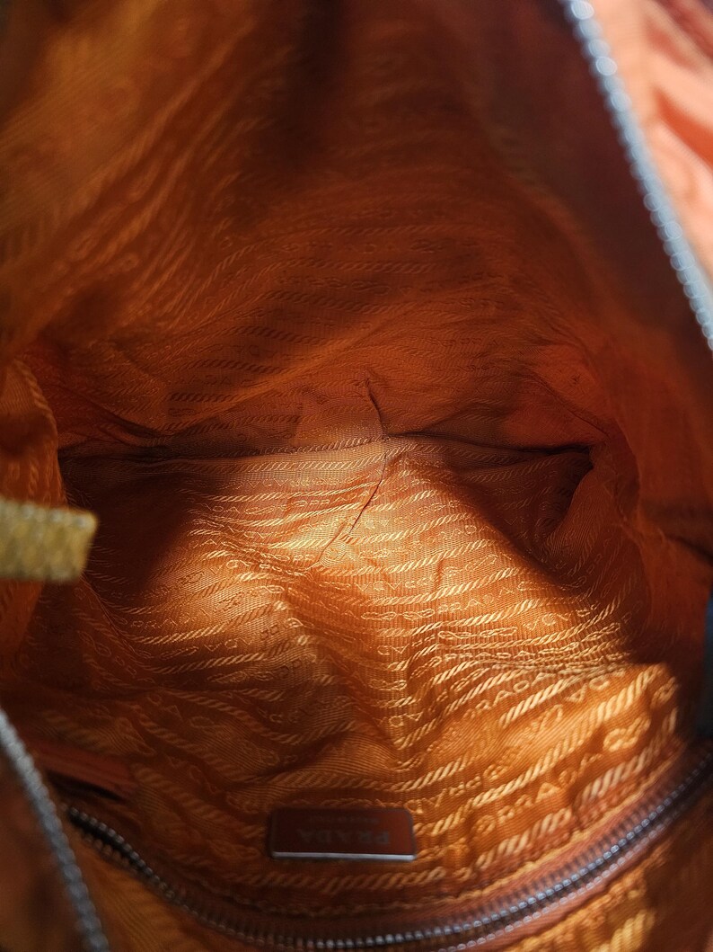 Peut inclure: Vue int&eacute;rieure d'un sac &agrave; main orange. La doublure est d'une nuance plus claire d'orange avec un motif r&eacute;p&eacute;titif. Une petite &eacute;tiquette m&eacute;tallique rectangulaire avec le nom de la marque est visible en bas. L'ext&eacute;rieur du sac est orange fonc&eacute;.