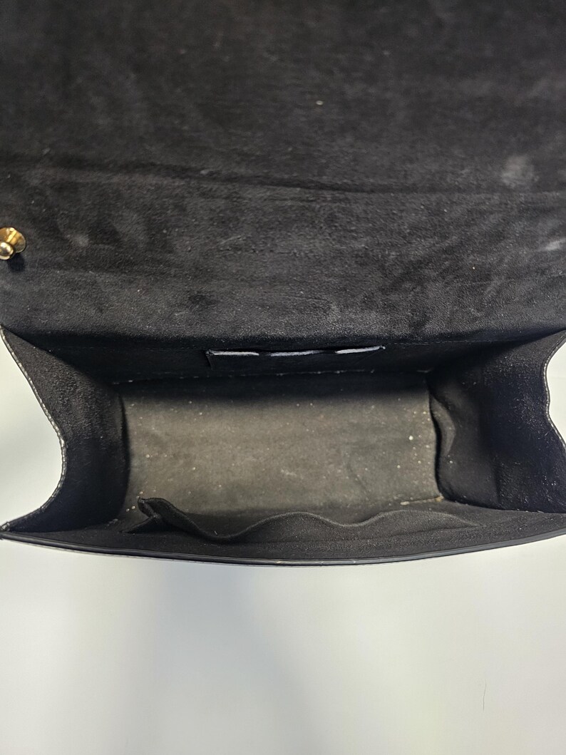 K&ouml;nnte beinhalten: Offene Ansicht des Inneren einer schwarzen Handtasche. Das Innere der Tasche ist mit schwarzem Stoff gef&uuml;ttert und hat eine kleine Tasche. Die Klappe der Tasche ist ge&ouml;ffnet und gibt den Blick ins Innere frei. Ein goldfarbener Verschluss ist sichtbar.