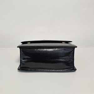 K&ouml;nnte beinhalten: Eine schwarze, rechteckige Handtasche mit gl&auml;nzendem Finish. Die Tasche hat einen Klappverschluss mit einem goldfarbenen Verschluss. Das Material hat ein strukturiertes, reptilienartiges Muster. Die Tasche steht vor einem schlichten wei&szlig;en Hintergrund.