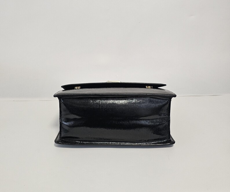 K&ouml;nnte beinhalten: Eine schwarze, rechteckige Handtasche mit gl&auml;nzendem Finish. Die Tasche hat einen Klappverschluss mit einem goldfarbenen Verschluss. Das Material hat ein strukturiertes, reptilienartiges Muster. Die Tasche steht vor einem schlichten wei&szlig;en Hintergrund.