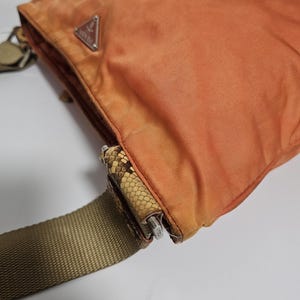 Peut inclure: Sac bandouli&egrave;re en nylon orange avec une bandouli&egrave;re r&eacute;glable beige. Le sac pr&eacute;sente un d&eacute;tail &agrave; motif peau de serpent et une fermeture &eacute;clair argent&eacute;e. Un logo triangulaire argent&eacute; est visible &agrave; l'avant du sac.