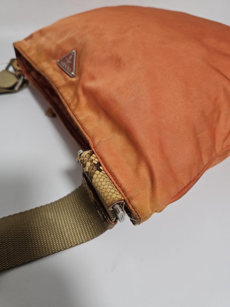Peut inclure: Sac bandouli&egrave;re en nylon orange avec une bandouli&egrave;re r&eacute;glable beige. Le sac pr&eacute;sente un d&eacute;tail &agrave; motif peau de serpent et une fermeture &eacute;clair argent&eacute;e. Un logo triangulaire argent&eacute; est visible &agrave; l'avant du sac.