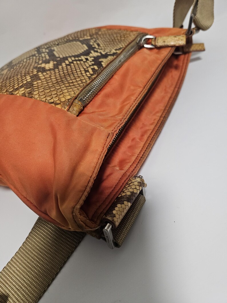 Peut inclure: Un sac bandouli&egrave;re orange avec un rabat en peau de serpent et une fermeture &eacute;clair argent&eacute;e. Le sac est dot&eacute; d'une bandouli&egrave;re tiss&eacute;e marron et beige. Le sac est fait de cuir et de tissu.