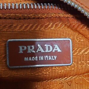 Peut inclure: Gros plan sur l'int&eacute;rieur d'un sac &agrave; main Prada orange. L'image montre le logo de la marque sur une &eacute;tiquette rectangulaire brun rouge&acirc;tre qui indique "PRADA MADE IN ITALY". Une fermeture &eacute;clair argent&eacute;e est visible en haut.