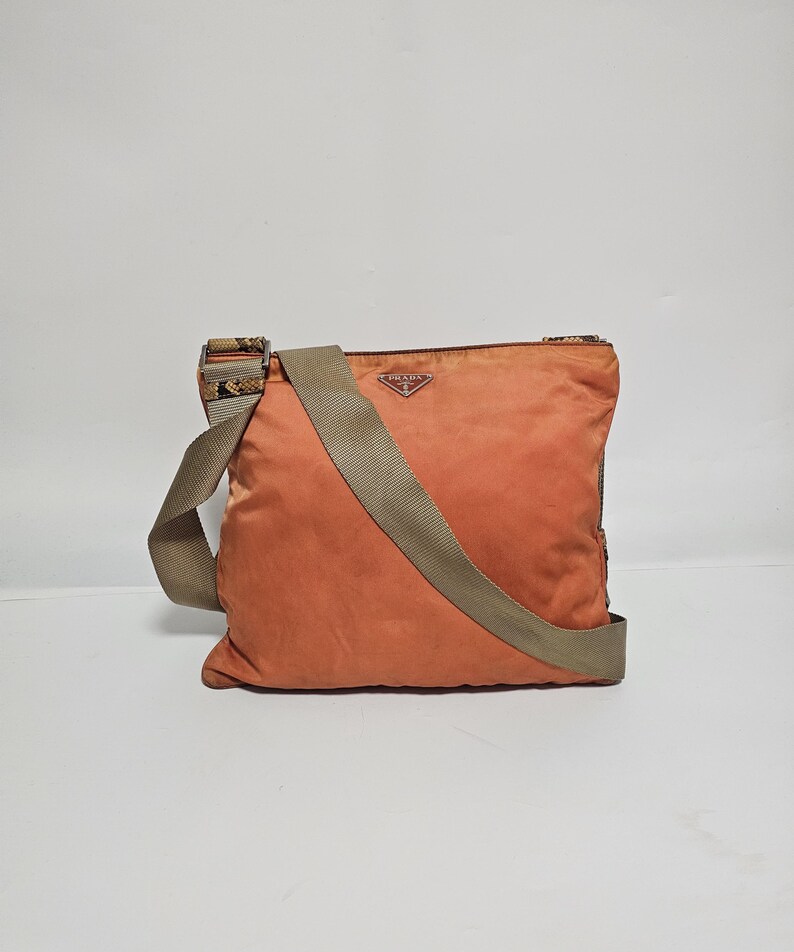 Peut inclure: Sac bandouli&egrave;re Prada orange avec une bandouli&egrave;re beige. Le sac est de forme carr&eacute;e et porte un petit logo Prada. La bandouli&egrave;re pr&eacute;sente un motif sur les extr&eacute;mit&eacute;s.