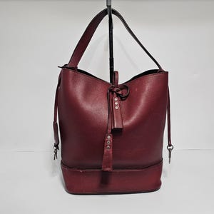 以下が含まれることがあります： 引き紐とタッセルが付いた、ワインレッドの革製のバケットバッグ。バッグには、シルバーのジッパーとシルバーのバックルが付いています。