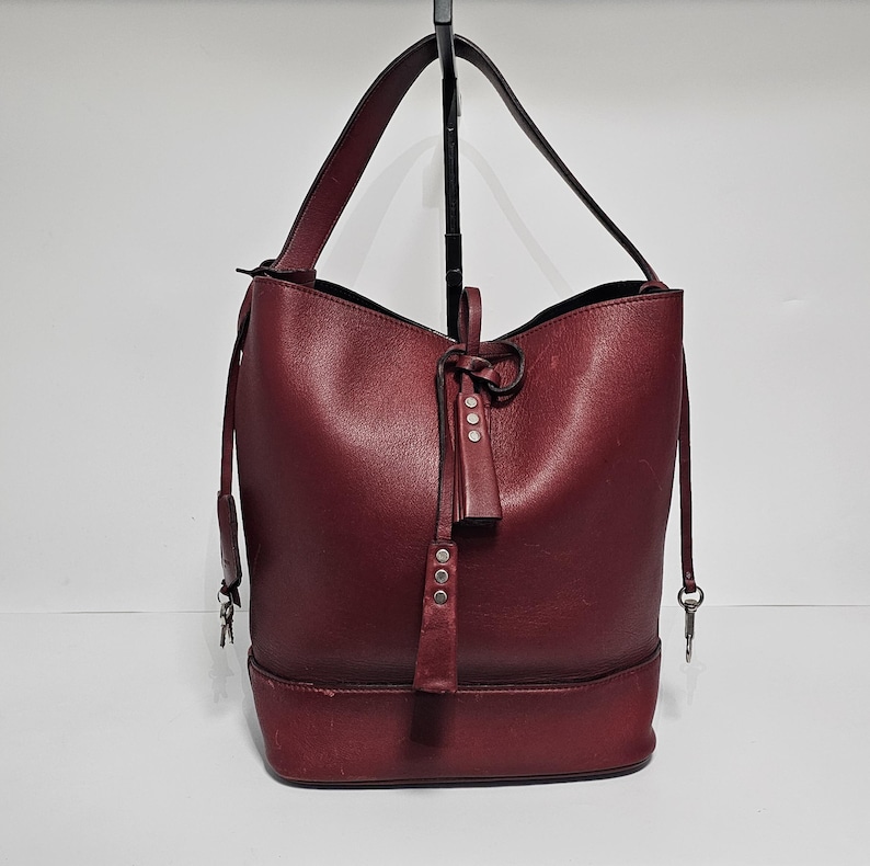 以下が含まれることがあります： 引き紐とタッセルが付いた、ワインレッドの革製のバケットバッグ。バッグには、シルバーのジッパーとシルバーのバックルが付いています。