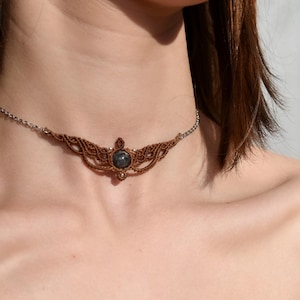 Peut inclure: Un collier ras du cou en macramé marron avec une pierre centrale gris foncé. Le collier présente un design délicat et tissé et une chaîne argentée. Le choker est autour du cou.