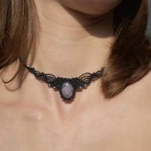 May include: Black macrame choker necklace with a central oval-shaped, light purple gemstone. The necklace features intricate knotting and small silver-colored beads. A stylish and unique piece of jewelry.