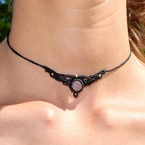 May include: Black macrame choker necklace with a central, round, light pink gemstone. The necklace features intricate knotting and small gold-colored beads. The choker is worn around the neck.