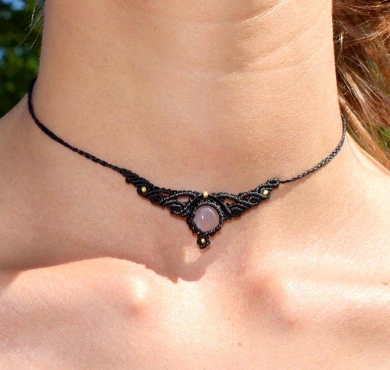 May include: Black macrame choker necklace with a central, round, light pink gemstone. The necklace features intricate knotting and small gold-colored beads. The choker is worn around the neck.