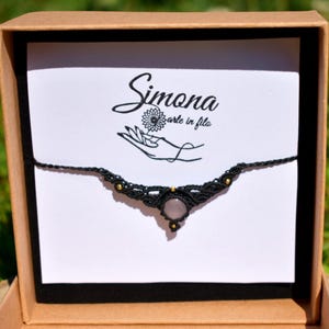 May include: A black macrame necklace with a central rose quartz stone and gold-colored beads, presented in a brown gift box. The card inside the box features the text "Simona arte in fila" and a hand-drawn logo.