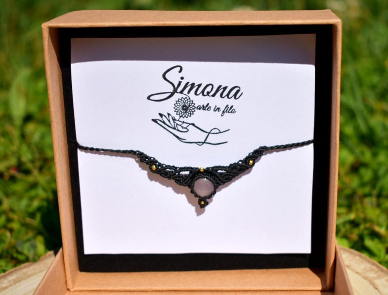 May include: A black macrame necklace with a central rose quartz stone and gold-colored beads, presented in a brown gift box. The card inside the box features the text "Simona arte in fila" and a hand-drawn logo.