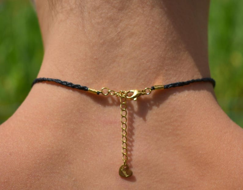 May include: Close-up of a black braided necklace with gold-colored clasp and extender chain. The necklace has a small heart-shaped charm at the end of the extender chain. The background is blurred, with green and brown tones.