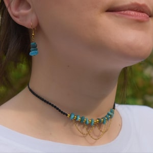 May include: A black cord choker necklace featuring a row of small, irregular turquoise-colored stone beads interspersed with gold-toned metal beads and delicate gold-toned chain drapes, paired with a matching single drop earring.