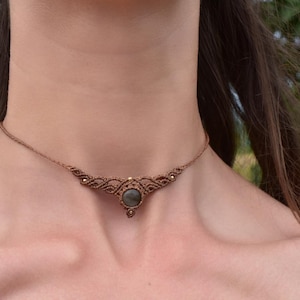 May include: A brown macrame choker necklace featuring a central round grey stone. The cord is intricately knotted in a symmetrical pattern that extends outward from the stone, with small metallic beads integrated into the woven design.