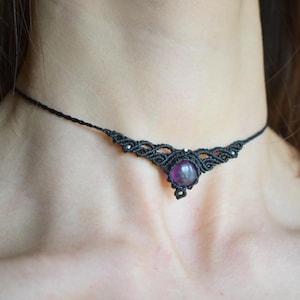 May include: Black macrame choker necklace with a central, round, purple gemstone. The choker features intricate knotting details and a delicate design. The necklace is worn around the neck.