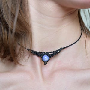 Può includere: Una collana girocollo in macramè nero con una gemma blu chiaro al centro. La collana presenta un'intricata annodatura e piccole perline color oro. Il girocollo è indossato intorno al collo.