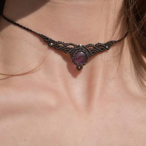 Può includere: Una collana girocollo in macramè nero con una gemma centrale rotonda e viola. La collana presenta un'intricata annodatura e piccole perline color argento. La collana è indossata intorno al collo.