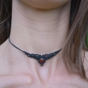 Peut inclure: Un collier ras-du-cou en macramé noir orné d'une pierre ronde rouge foncé en son centre. Le design complexe en cordon noué intègre de petites perles métalliques dans le motif, créant une pièce décorative symétrique qui se porte près du cou.