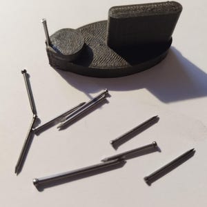 Può includere: Una base in plastica grigia stampata in 3D con una piccola piattaforma rotonda e una piattaforma rettangolare. Ci sono diversi chiodi di metallo argentati sparsi intorno alla base.